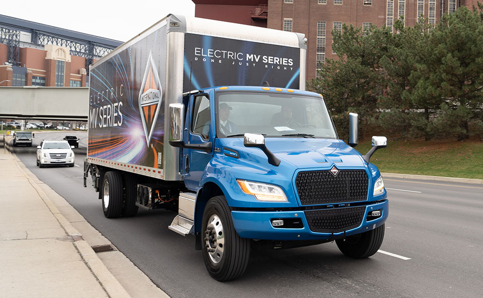 Small, white dry freight body with blue decals, mounted on Isuzu NPR electric chassis, photographed from behind while driving on a highway